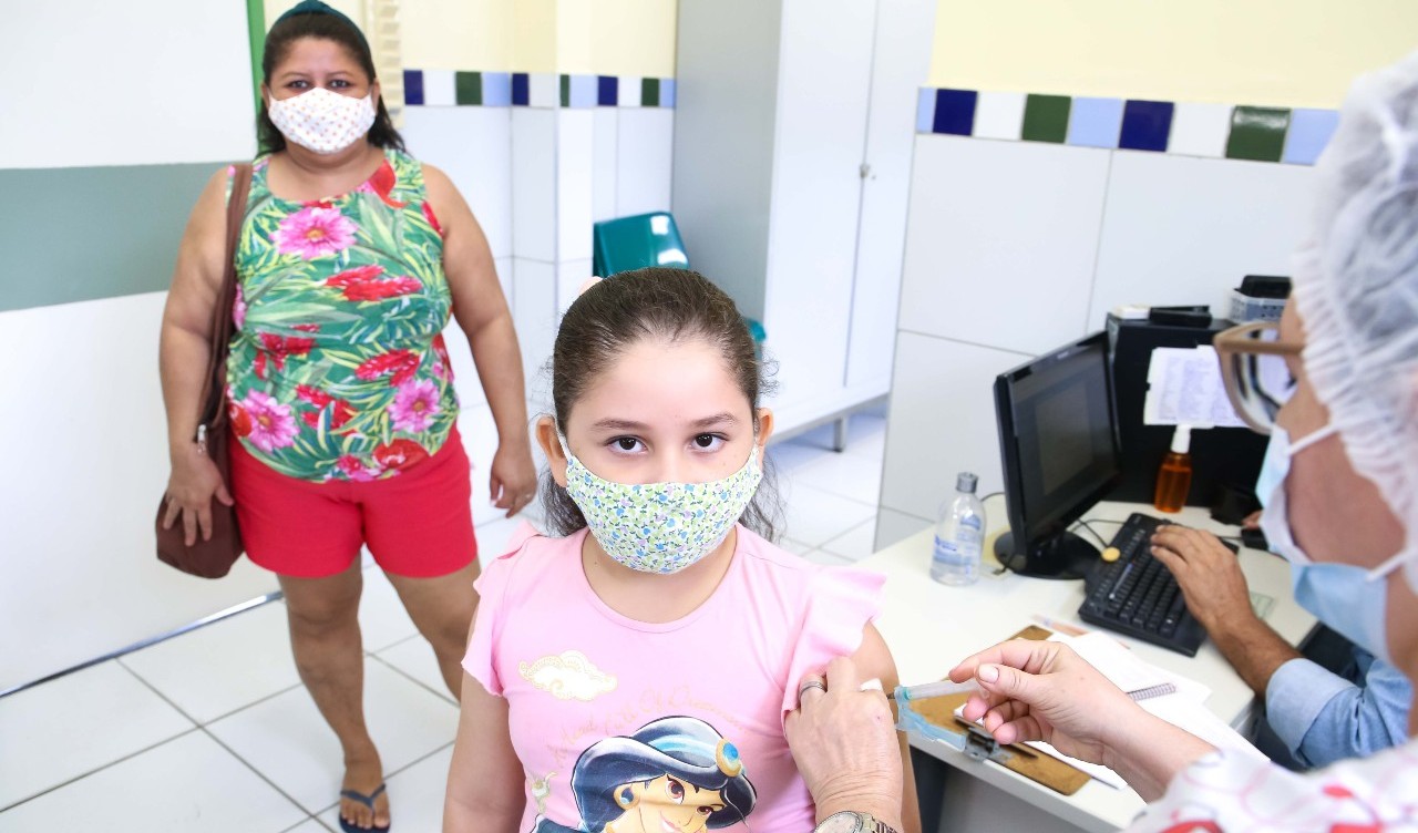 em primeiro plano, uma menina de máscara é vacinada por uma enfermeira. ao fundo, a mãe dela, em pé, observa